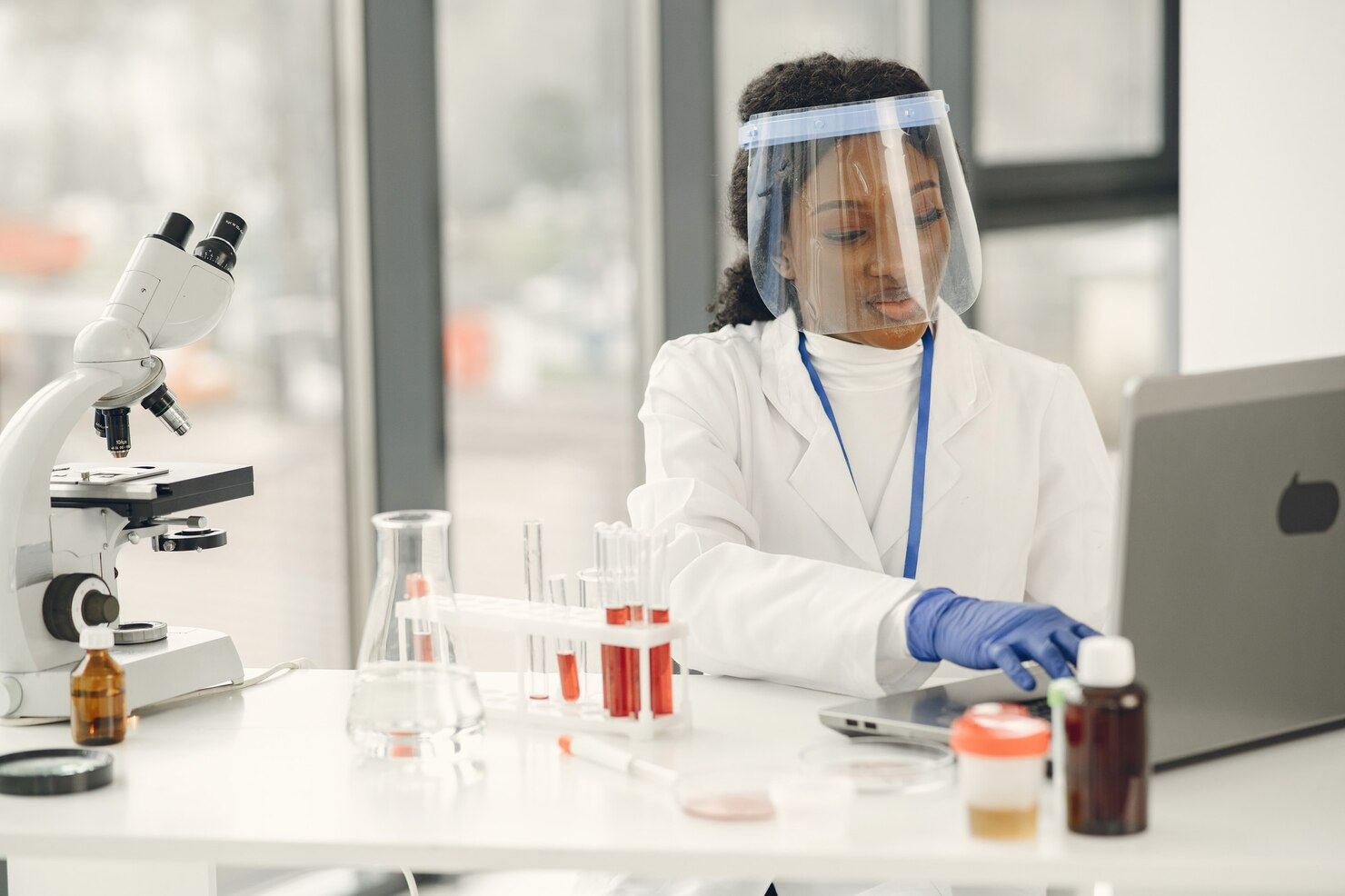 African scientist in lab coat and face shield working with test tubes and a laptop